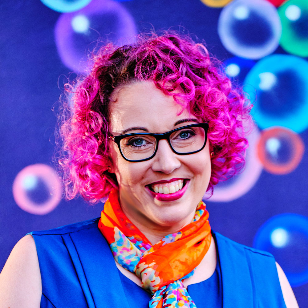 A photo of Jo Minney with bright pink curly hair and dorky black glasses. Jo is wearing an orange scarf and standing in front of a mural of bubbles.